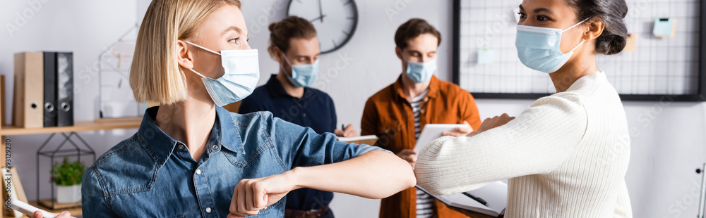 young businesswomen in medical masks doing elbow bump near colleagues on blurred background, banner young businesswomen in medical masks doing elbow bump near colleagues on blurred background, banner