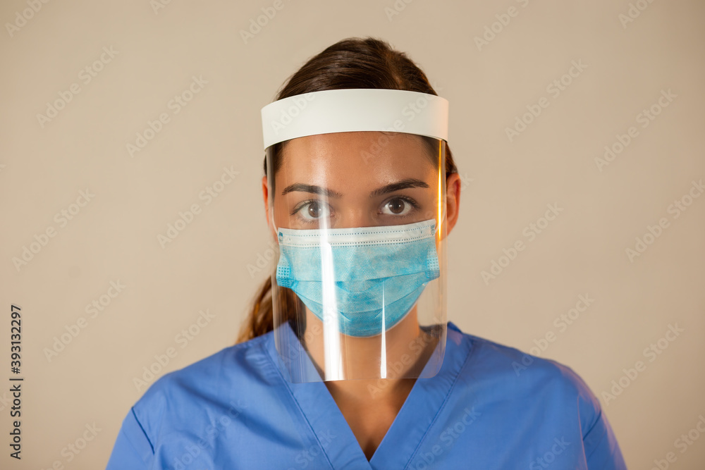 Caucasian female with attached face shield with light background. Medical worker with protective equipment looking into camera. Female in blue shirt with face mask on. Caucasian female with attached face shield with light background. Medical worker with protective equipment looking into camera. Female in blue shirt with face mask on.