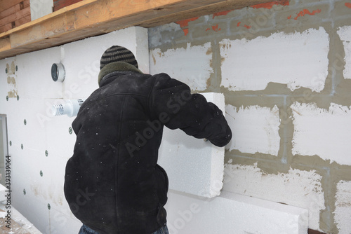 A building contractor is installing an external solid wall insulation fixing insulating polystyrene panels to the wall with ventilation duct constructed from autoclaved aerated concrete blocks.