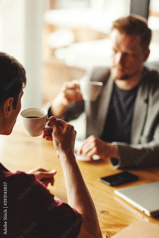 Over the shoulder view of young businessman having informal meeting over coffee with defocused middle aged colleague sittign at table in cafe Over the shoulder view of young businessman having informal meeting over coffee with defocused middle aged colleague sittign at table in cafe