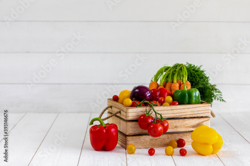 Une caisse de légumes fraiche avec des tomates, des oignons,des poivrons,des carottes,des tomates cerises avec de belles couleurs éclatantes rouges, jaunes,vert et oranges sur un fond de bardage blanc