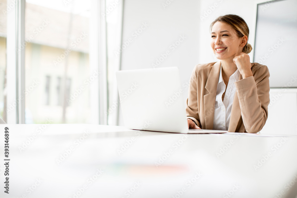 Young woman working on laptop in bright office with big screen behind her Young woman working on laptop in bright office with big screen behind her