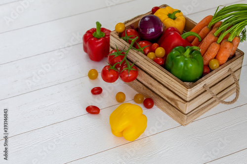 Une caisse de légumes fraiche avec des tomates, des oignons,des poivrons,des carottes,des tomates cerises avec de belles couleurs éclatantes rouges, jaunes,vert et oranges sur un fond de bardage blanc