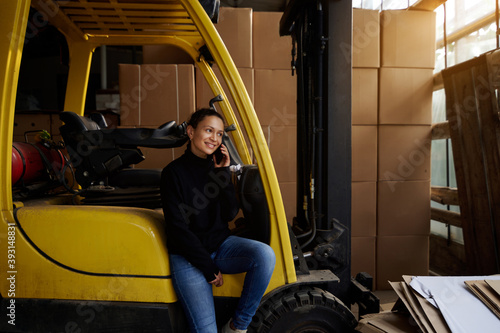 Inside the warehouse, a young woman businessman sits on the step of a yellow forklift and speaks on the phone. The sun is shining through the warehouse windows.