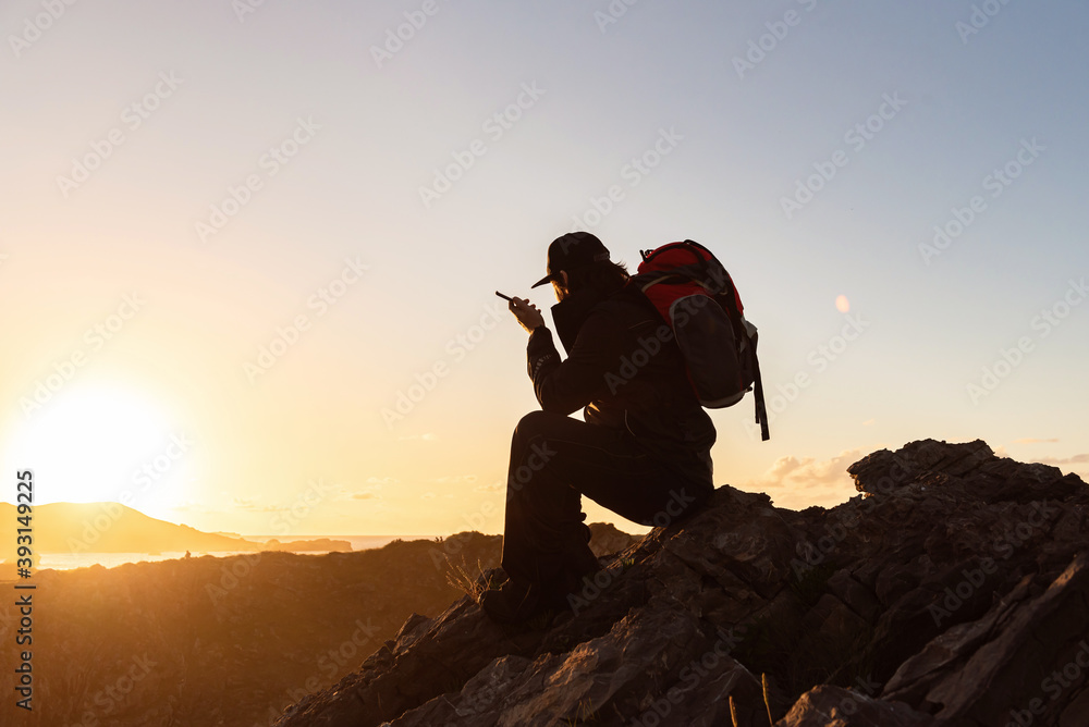 Side view of unrecognizable male hiker sitting on rock in highlands and speaking on cellphone while admiring sunset Side view of unrecognizable male hiker sitting on rock in highlands and speaking on cellphone while admiring sunset