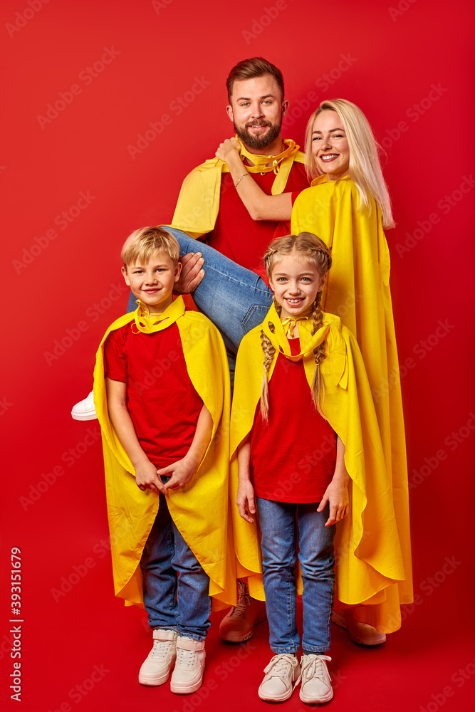 good-looking parents playing superheroes with children, posing isolated over red background, wearing yellow cloaks
