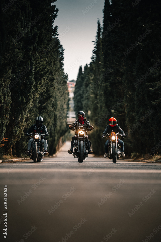 Group of bikers in black leather jackets and helmets riding powerful motorcycles on asphalt road leading between green forest in countryside Group of bikers in black leather jackets and helmets riding powerful motorcycles on asphalt road leading between green forest in countryside