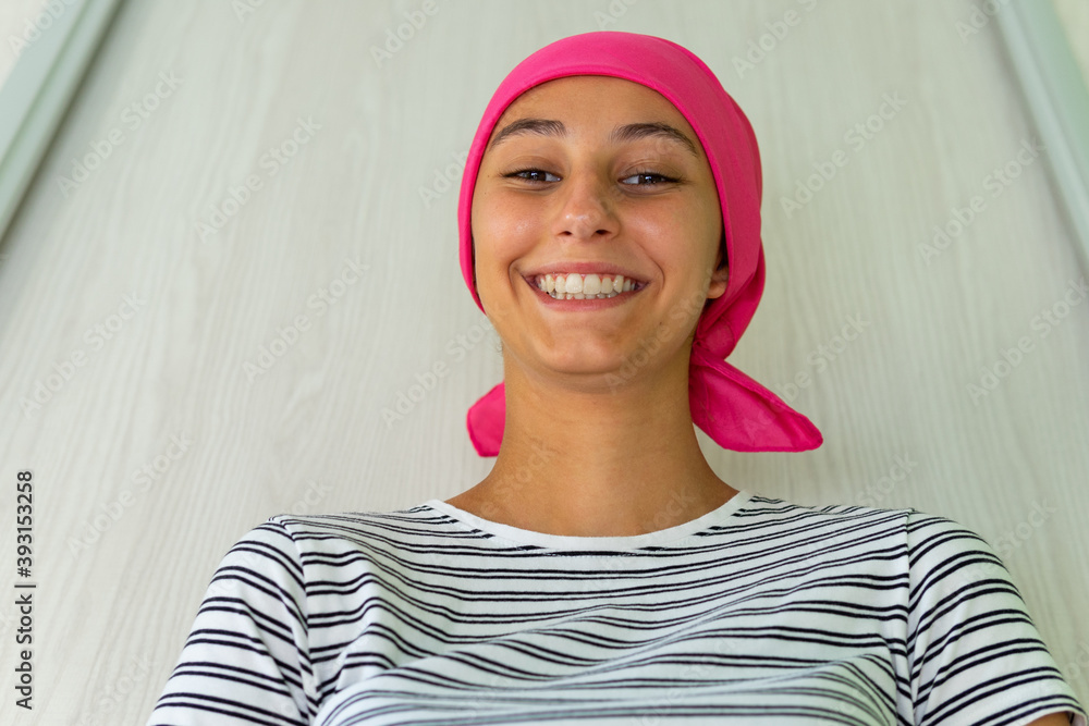 Young delighted female with cancer wearing headscarf standing in apartment and looking at camera Young delighted female with cancer wearing headscarf standing in apartment and looking at camera