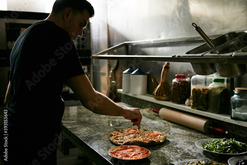 Side view of male pizza maker cutting freshly baked appetizing hot pizza on marble counter while working in restaurant kitchen photo