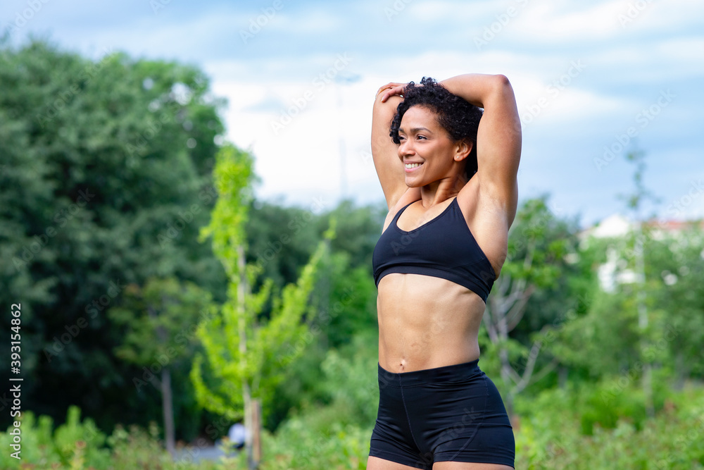 Delighted African American muscular female stretching arms and warming up before training in park while looking away Delighted African American muscular female stretching arms and warming up before training in park while looking away