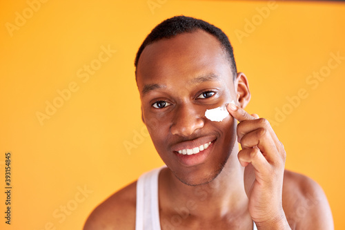 young african-american guy applying face cream under his eyes on yellow background, he likes taking care of skin, daily procedure in the morning or before going to bed