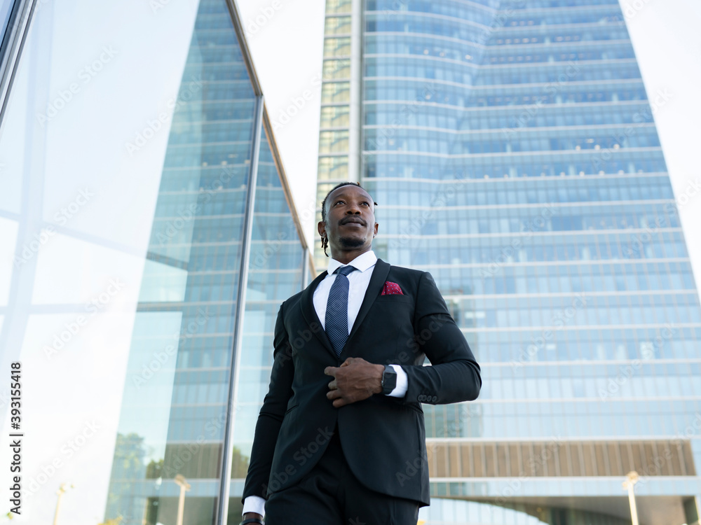 Serious African American male entrepreneur wearing elegant suit walking along glass office building in city center and looking away Serious African American male entrepreneur wearing elegant suit walking along glass office building in city center and looking away