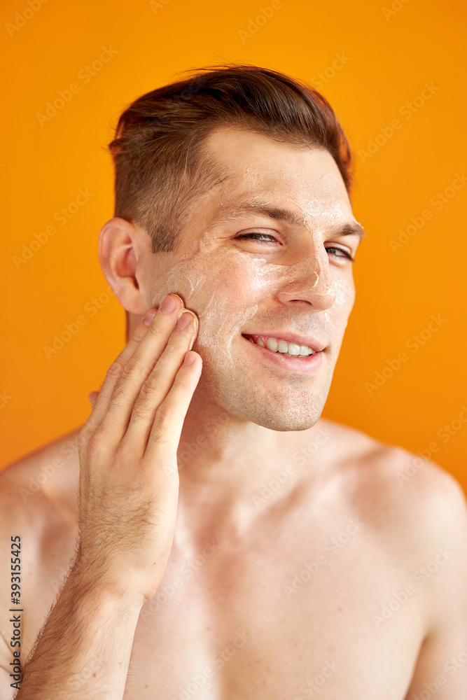 Obraz premium young caucasian man apply moisturizing face cream for healthy glowing skin, isolated over yellow background, handsome guy use nourishing facial balm, skincare, hygiene concept