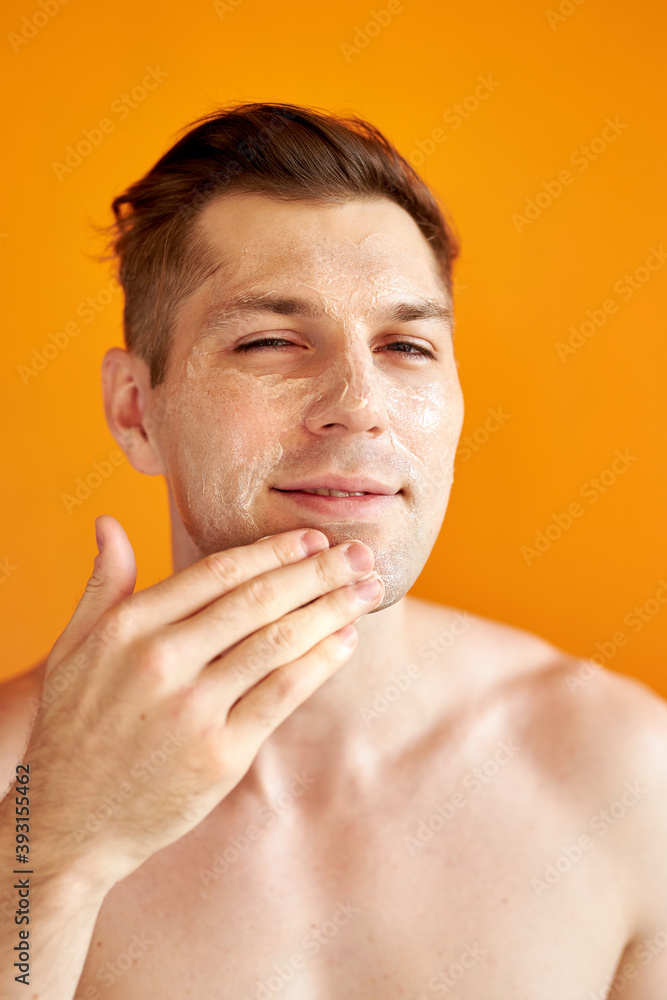 Fototapeta premium young caucasian man apply moisturizing face cream for healthy glowing skin, isolated over yellow background, handsome guy use nourishing facial balm, skincare, hygiene concept