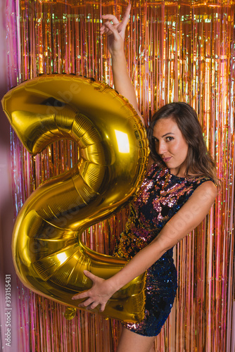 Side view of young female in bright festive outfit embracing golden balloon in shape of number 2 against colorful tinsel while celebrating New Year holiday photo