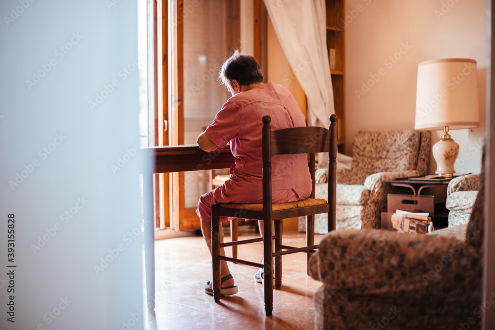 Back view of a senior woman with Alzheimer's mental health issues painting on a notebook inside her home Back view of a senior woman with Alzheimer's mental health issues painting on a notebook inside her home