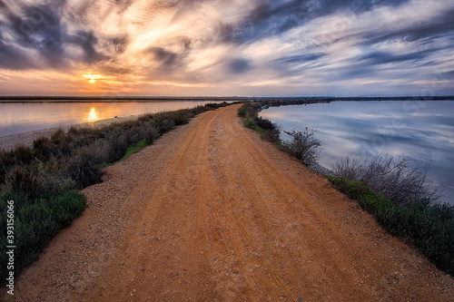 dirt road that divides the waters  on the left the sunset with orange colors and on the right clouds and blue tones