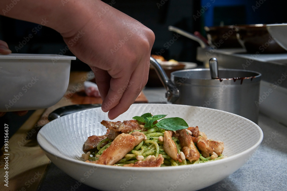 Closeup of crop anonymous cook seasoning and garnishing spaghetti with pesto sauce and chicken served in white bowl while working in restaurant kitchen Closeup of crop anonymous cook seasoning and garnishing spaghetti with pesto sauce and chicken served in white bowl while working in restaurant kitchen