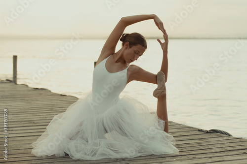 Full body flexible classic ballet female dancer in elegant white dress performing sensual pose on wooden pier against blurred sea in summer evening