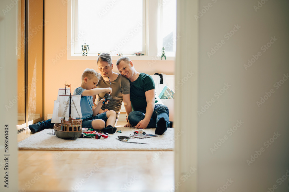 Homosexual fathers looking at child playing while making toy boat at home Homosexual fathers looking at child playing while making toy boat at home