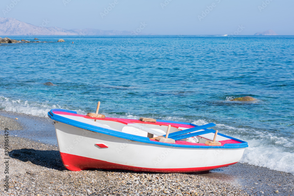Colorful painted wooden boat on the shore of a stony beach with calm blue water in Ikaria, Greece.