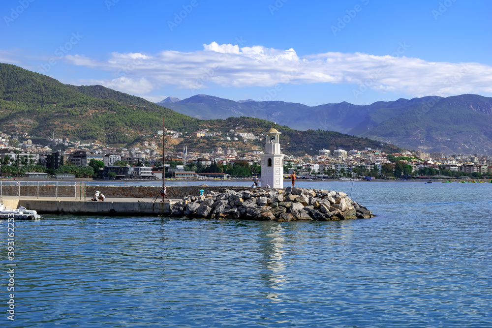 White Alanya lighthouse against the background of the panorama of the city - view from the Mediterranean Sea. Beautiful seascape from the embankment of the Turkish resort among the mountains