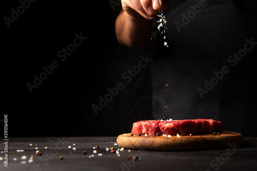 cook sprinkles salt on a piece of meat on a black background. The hand of the cook is in the frame. Horizontal photo with free space for text logos and advertisements on the left side.