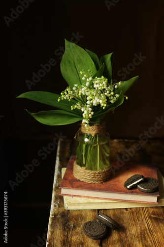 On a wooden background  a vase with a bouquet of white lilies of the valley standing on books and chocolate milk. 