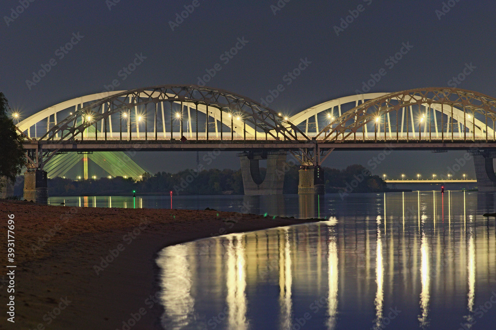 Three illuminated bridges at night. Picturesque landscape of Dnipro River with arched Railway bridge, Darnytskyi Rail and road Bridge. Pivdennyi Bridge in the background. Scenic evening landscape