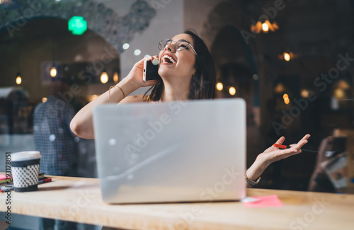 Toothy Caucaisan woman in optical eyewear laughing during funny cellphone conversation, cheerful female freelancer with netbook feeling delight happiness during friendly smartphone call in coworking