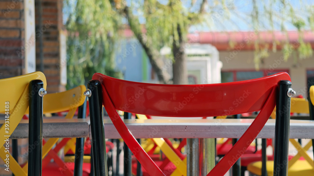 Tables and metal chairs with vivid red and yellow colors in harmony with green trees and blue sky in an open air restaurant at lunch time in a university campus, empty because of the Covid-19 pandemic