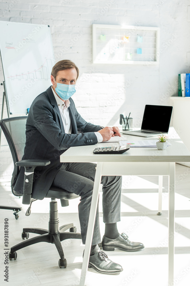 Full length of businessman in medical mask looking at camera, while writing in notebook, sitting at workplace in office Full length of businessman in medical mask looking at camera, while writing in notebook, sitting at workplace in office
