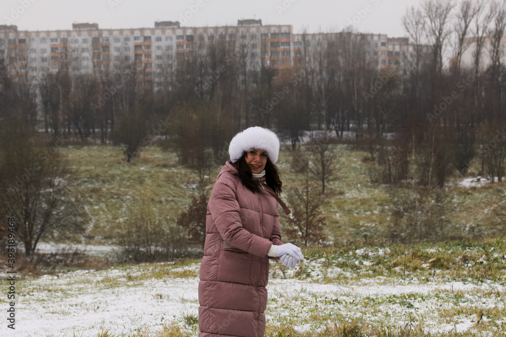 Fototapeta premium A girl in winter clothes on a walk in the park. Sculpts a ball of snow with his hands. The ground is covered with the first snow