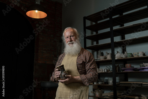 Mature craftsman in apron holding clay bowl. He is posing for camera in workshop. Concept of ceramic art and hobby. Old male potter