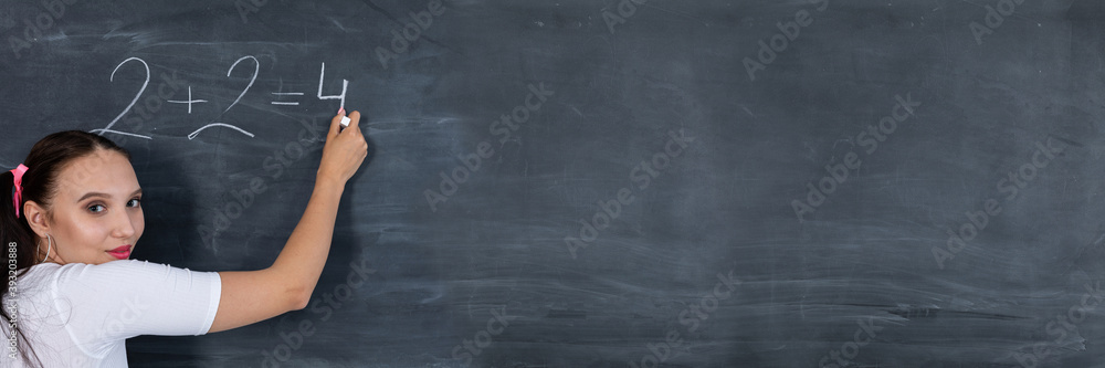 Panoramic view as a teenage girl chalks a math equation on a dark gray school blackboard. Her hair is pinned with a pink ribbon in two ponytails.