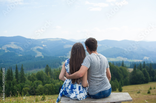 Back view of Romantic young couple enjoy the view of the mountains sitting on a bench on a sunny day in Carpatians. Loving couples date. Romantic outdoors.