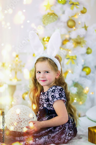 A beautiful little girl in a rabbit costume holds her finger in front of her lips on the background of a Christmas tree on Christmas day.