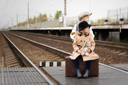 A little boy and a girl are sad on the platform near the railway tracks. Dressed in old clothes. Girl sitting on an old suitcase
