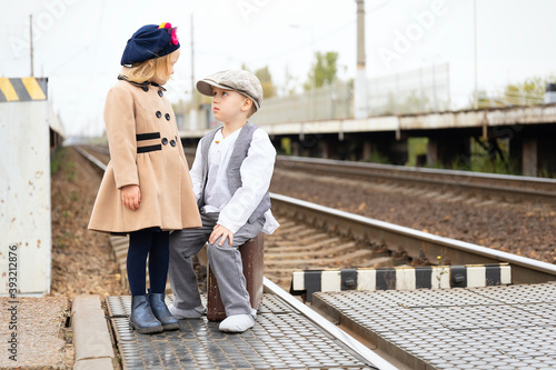 Little boy and girl in retro clothes on the train tracks at the station. The boy is not sitting in an old suitcase. They look at each other. © zerbob2