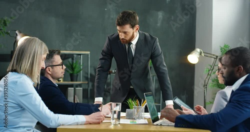 Caucasian handsome businessman standing at table with multiethnical business partners and talking about investment and project. Male boss leading and rulling at meeting of company mixed-races staff. photo