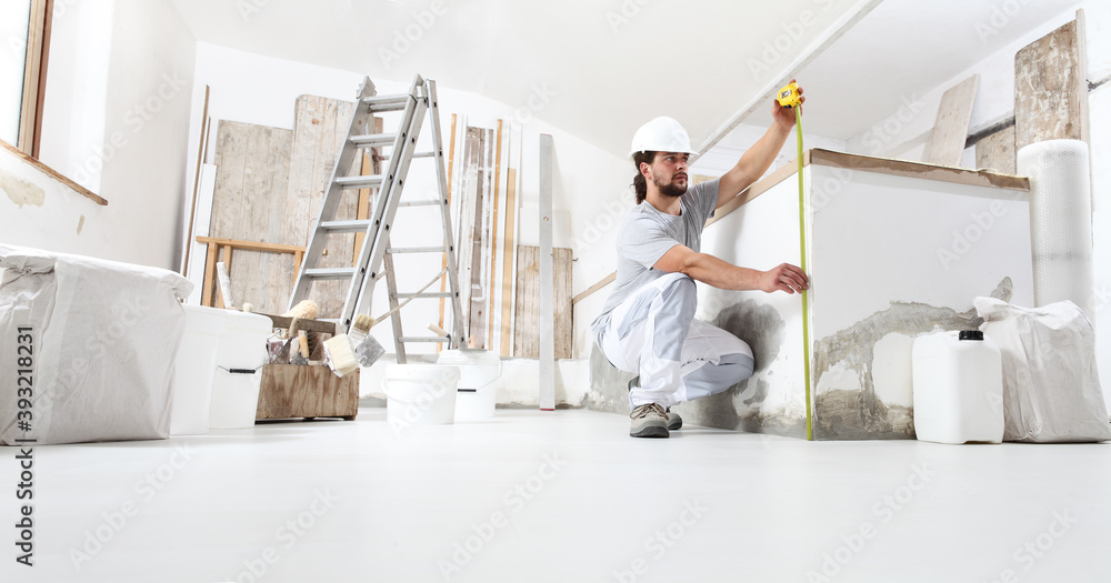 construction worker plasterer man measuring wall with measure tape in building site of home renovation with tools and building materials on the floor