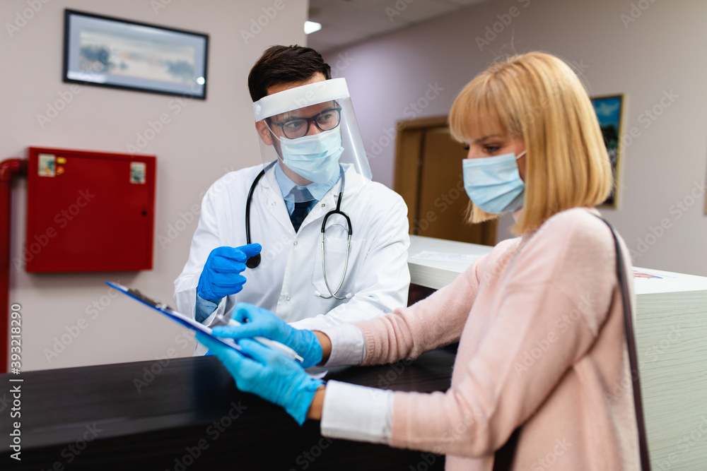 Young male practitioner with face shield and gloves working at clinic reception desk and talking with patient or visitor. Young male practitioner with face shield and gloves working at clinic reception desk and talking with patient or visitor.