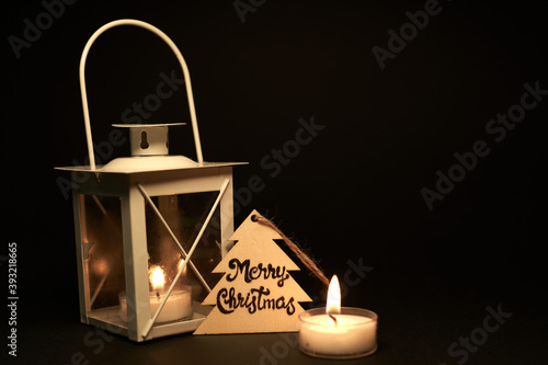 white candlestick with candles and christmas tree shaped christmas card on black background. Merry Christmas. christmas message on piece of wood lit by candles