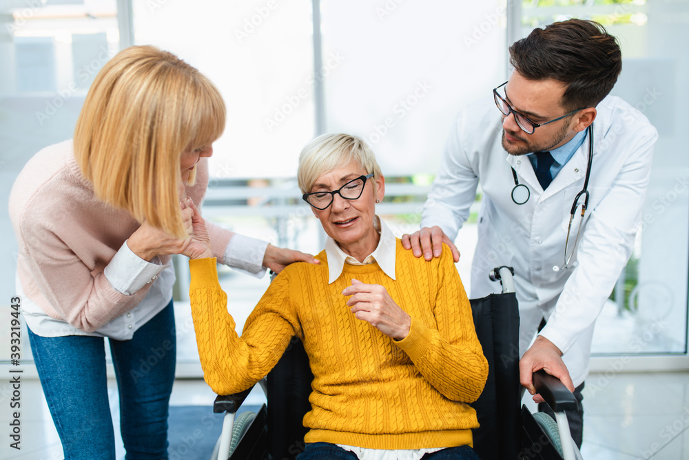 Fototapeta premium Young male practitioner working at medical clinic reception desk and helping to middle age woman and her sick mother which sitting in disability chair.