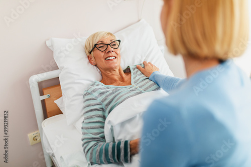 Beautiful middle age woman spending some time with her mother in hospital or nursing center. Senior woman lying in bed.