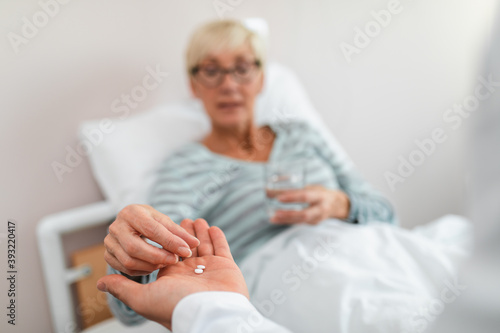 Young doctor visiting sick senior woman in hospital room or nursing center. Old woman also taking some pills from doctor's hand.