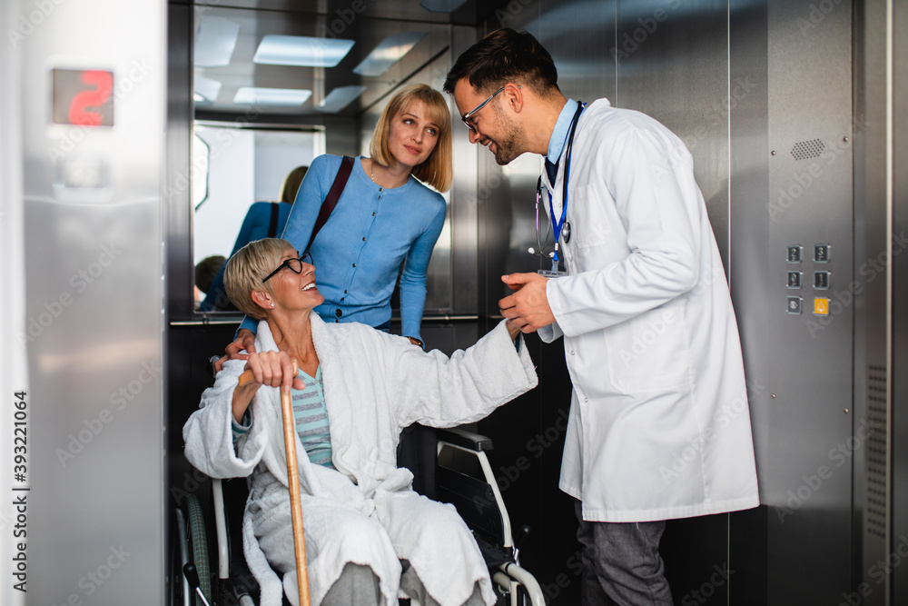 Young male practitioner talking with middle age woman and her sick mother which sitting in disability chair. They are standing in hospital or nursing center elevator. Young male practitioner talking with middle age woman and her sick mother which sitting in disability chair. They are standing in hospital or nursing center elevator.