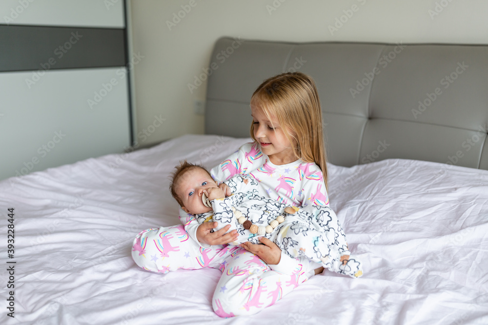 Little sister and her baby brother. Toddler kid playing with new sibling. Girl and baby 2-3 months old relax in a home bedroom. Family with two children at home. Love, trust and tenderness concept Little sister and her baby brother. Toddler kid playing with new sibling. Girl and baby 2-3 months old relax in a home bedroom. Family with two children at home. Love, trust and tenderness concept