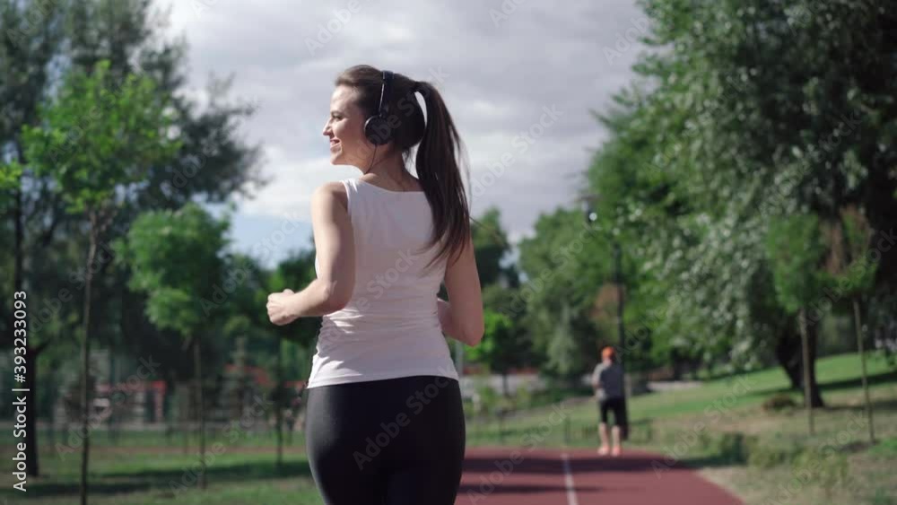Camera follows slim happy Caucasian sportswoman in headphones jogging on running track on summer morning. Back view of fit healthy woman exercising outdoors. Workout concept. Camera follows slim happy Caucasian sportswoman in headphones jogging on running track on summer morning. Back view of fit healthy woman exercising outdoors. Workout concept.