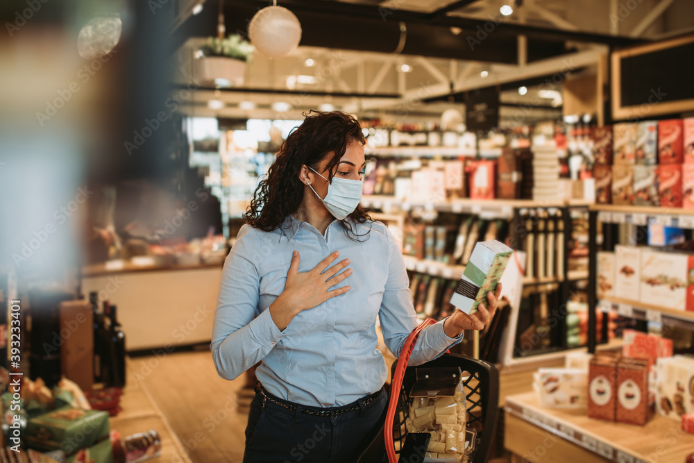 Beautiful young and elegant woman with face protective mask buying healthy food and drink in a modern supermarket or grocery store. Pandemic or epidemic lifestyle and consumerism concept.